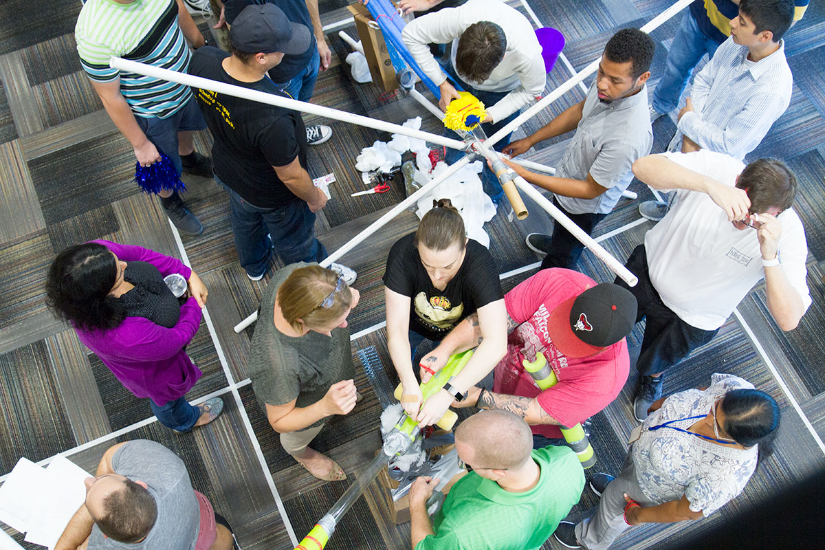 An overhead view of a dozen StrongMinders working on putting together PVC pipe with duct tape, pool noodles and string.